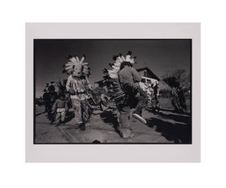 Black and white photograph of a circle of people dancing and wearing traditional Native America…