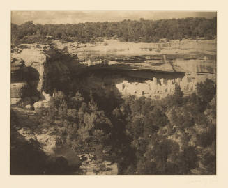 Greyscale photograph of a cliff dwelling in a canyon wall surrounded by trees 