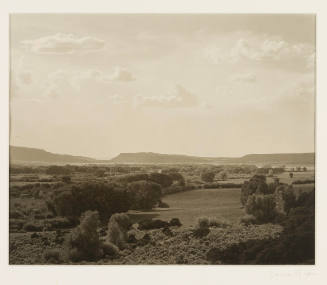 Greyscale photograph of a ranch with mesas in the background under a mostly clear sky