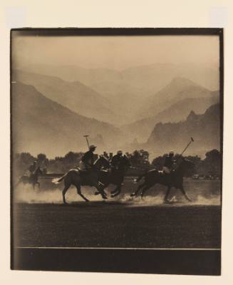 Greyscale photograph of a polo match with horses kicking up dust in front of mountains 