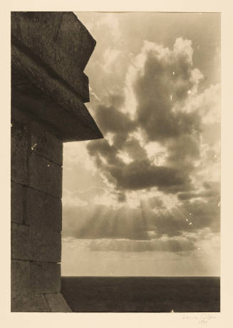 Greyscale photograph of a brick building's corner with the sun shining through clouds to the ri…