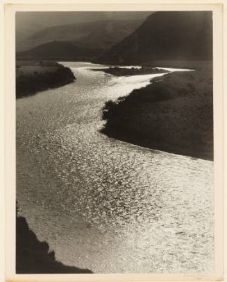 Greyscale photograph of a shimmering river with a small, distant island winding through a valle…